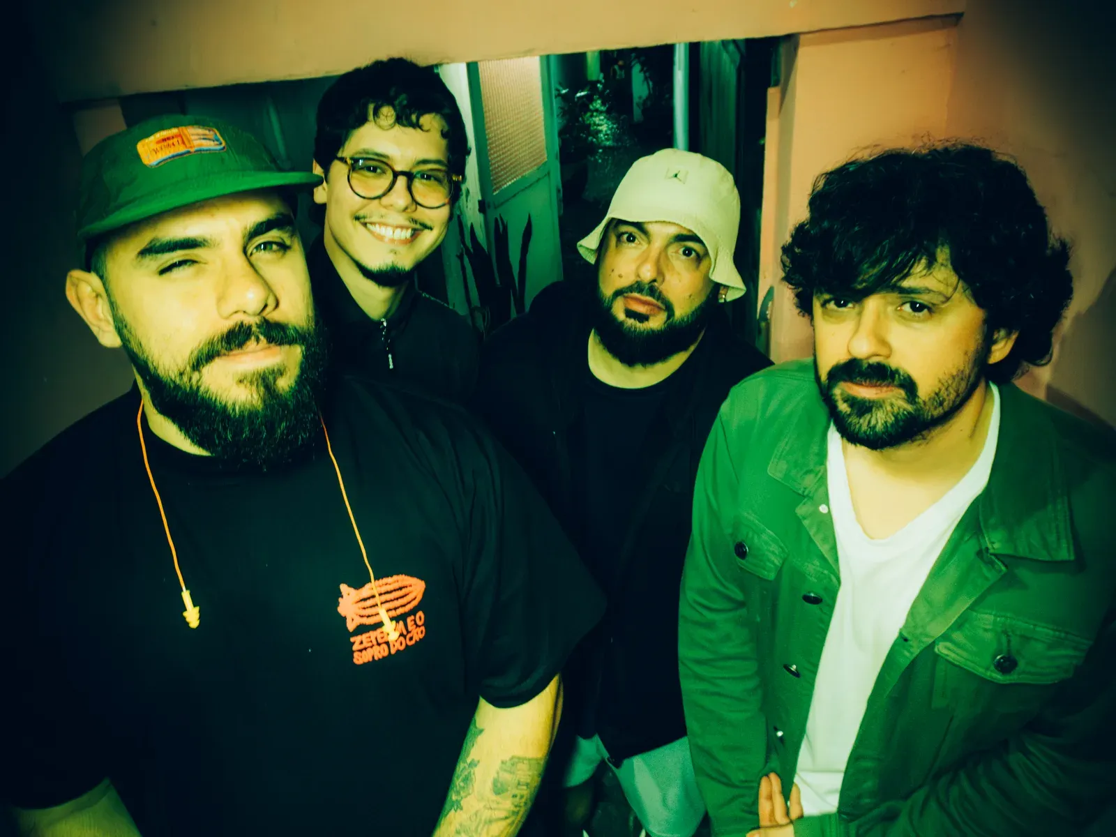 Os quatro integrantes da banda, vistos de cima, num corredor estreito de parede rosa sob luz verde