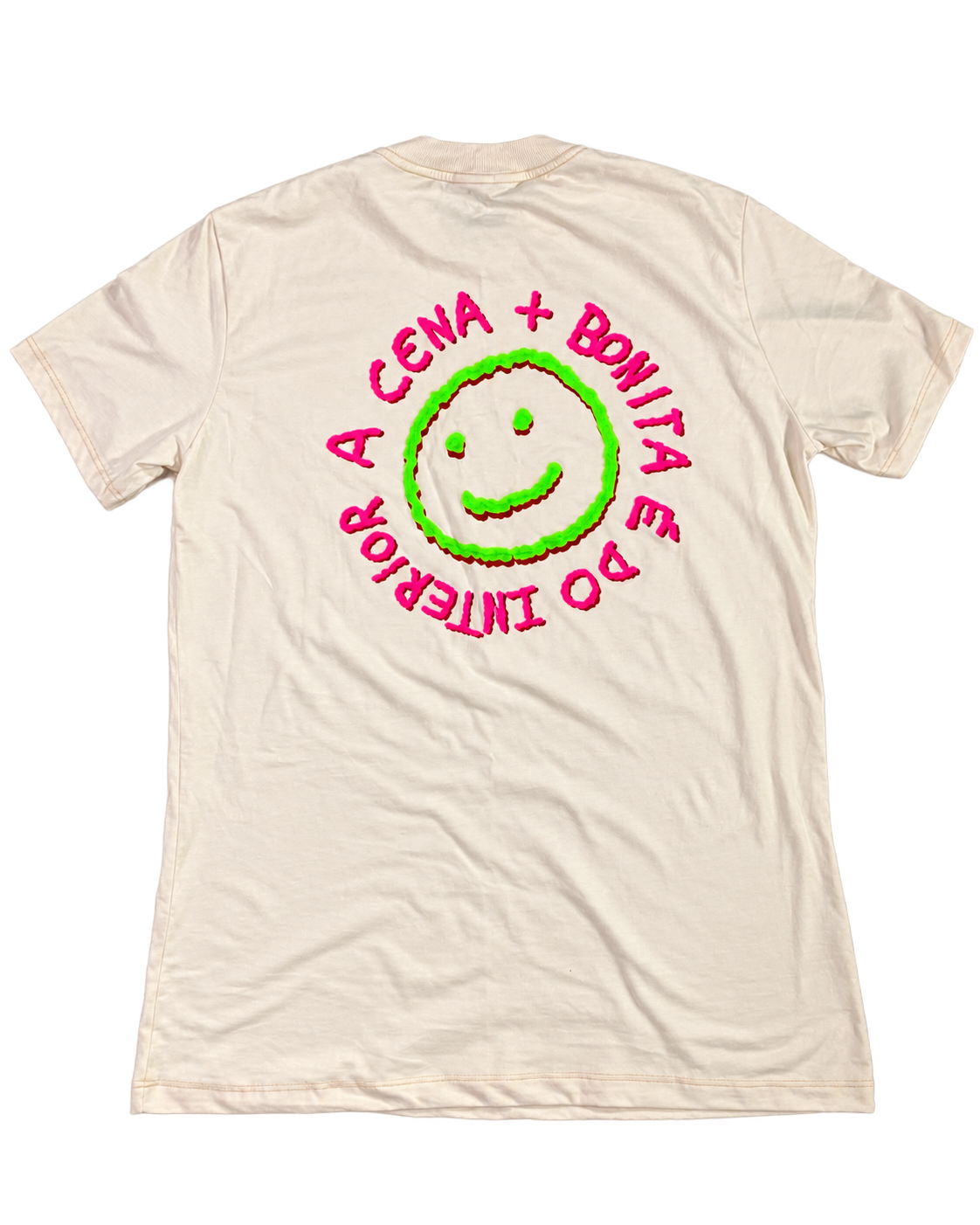 Camiseta creme com estampa nas costas: carinha sorridente verde e rosa cercada pela frase A cena + bonita é do interior